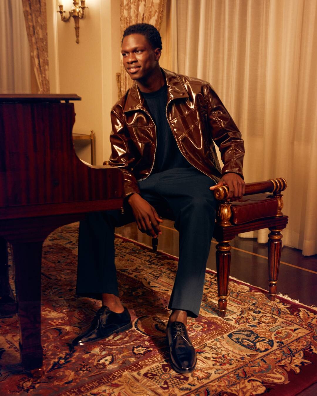 Man seated beside a piano, wearing a shiny brown jacket, dark pants, and classic shoes, in a sophisticated, elegant setting with a Persian rug and refined decor.