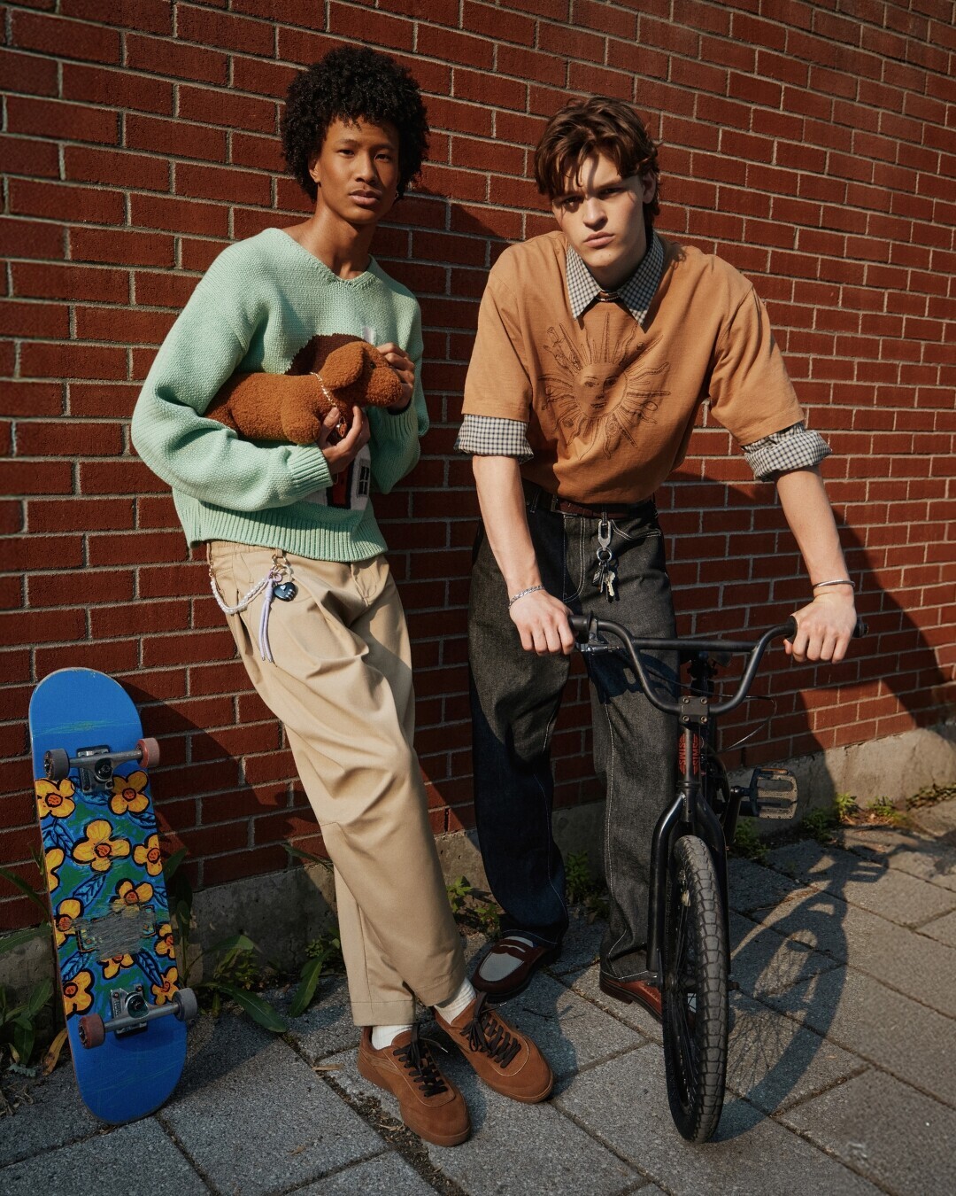 Man in a brown leather jacket, gray t-shirt, and light gray pants, crouching confidently and looking to the side, wearing casual sneakers.