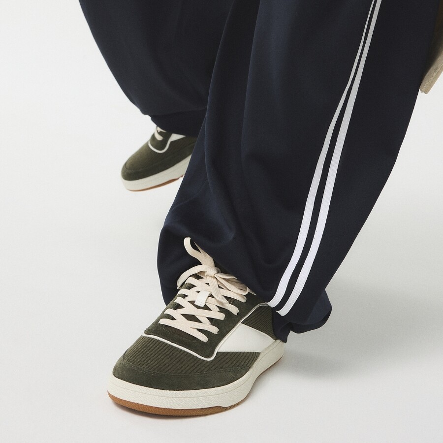 Man in a brown leather jacket, gray t-shirt, and light gray pants, crouching confidently and looking to the side, wearing casual sneakers.