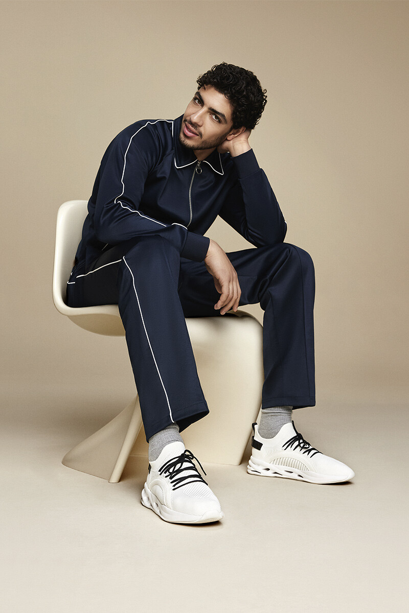 Man with slightly curly hair sitting on a beige chair, wearing a navy blue tracksuit, with one hand holding his head, and wearing white trainers with black laces.