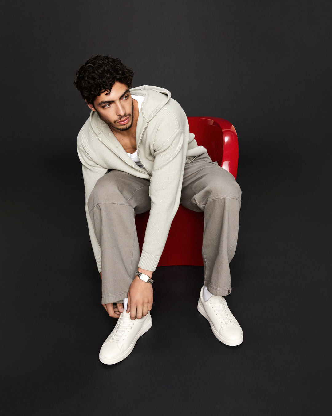 A stylish man ties his white sneakers while sitting on a red chair, dressed in a light hoodie and neutral-toned pants.