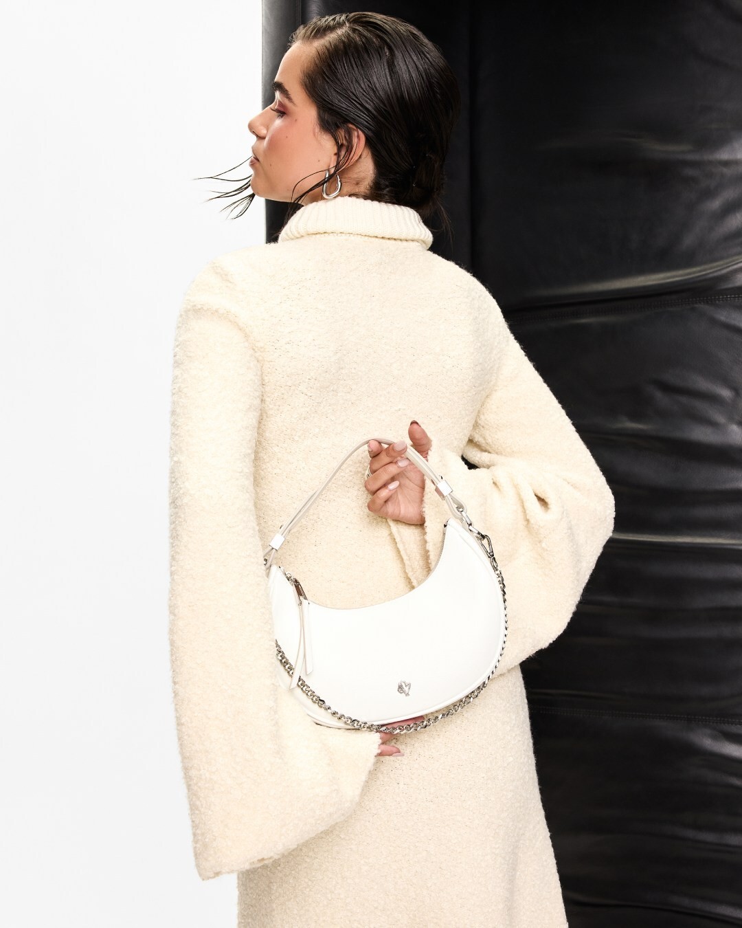 Woman in a cozy cream sweater dress holding a white shoulder bag with chain details behind her back, looking over her shoulder with a stylish, confident pose.