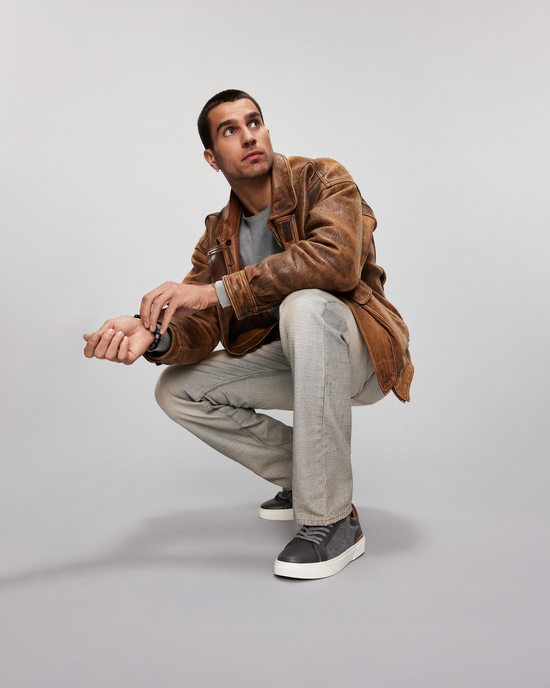 A man casually seated, wearing an oversized jacket, paired with grey jeans and dark grey sneakers.