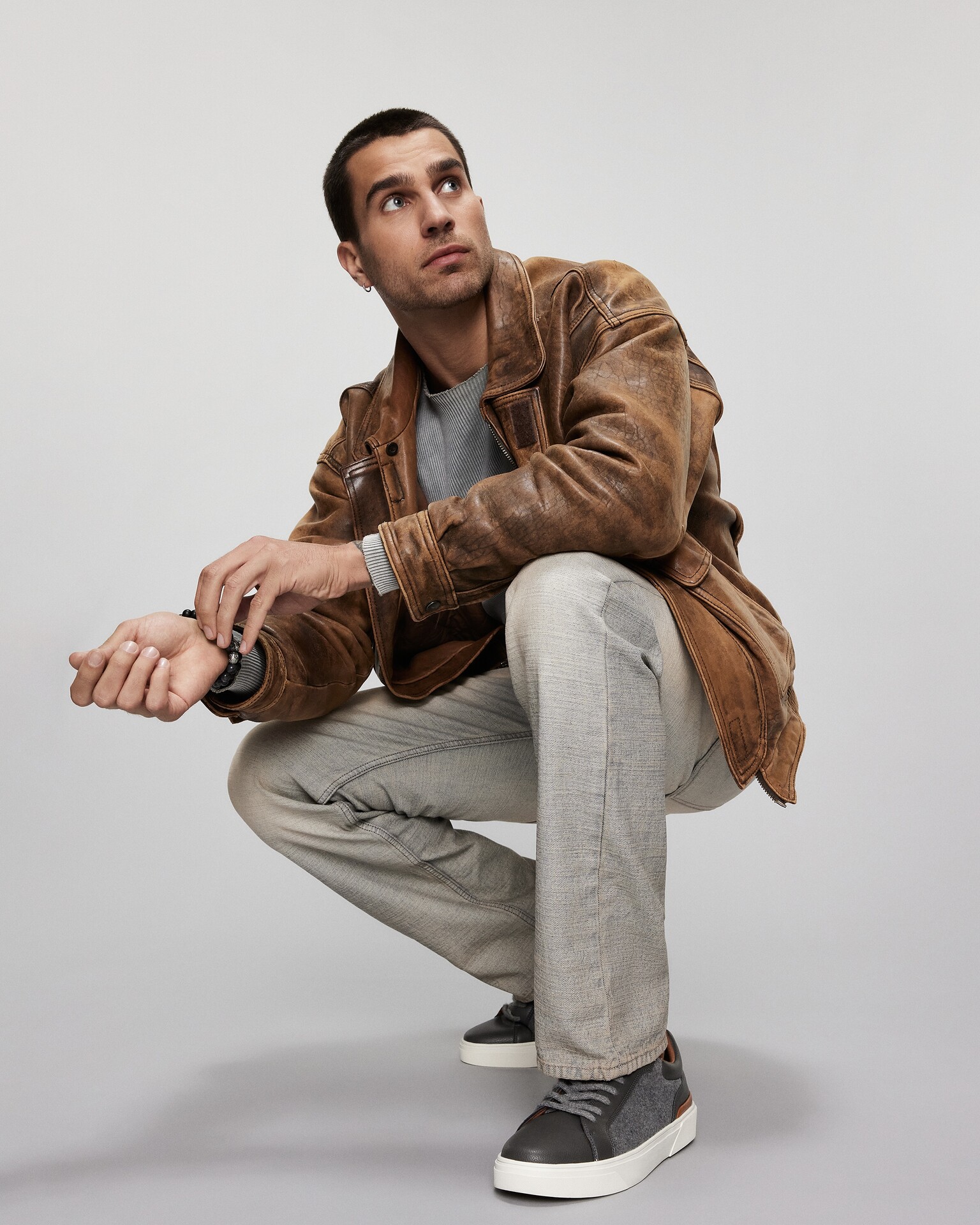 Man crouched down fiddling with his watch, wearing a brown coat, grey trousers and ALDO sneakers