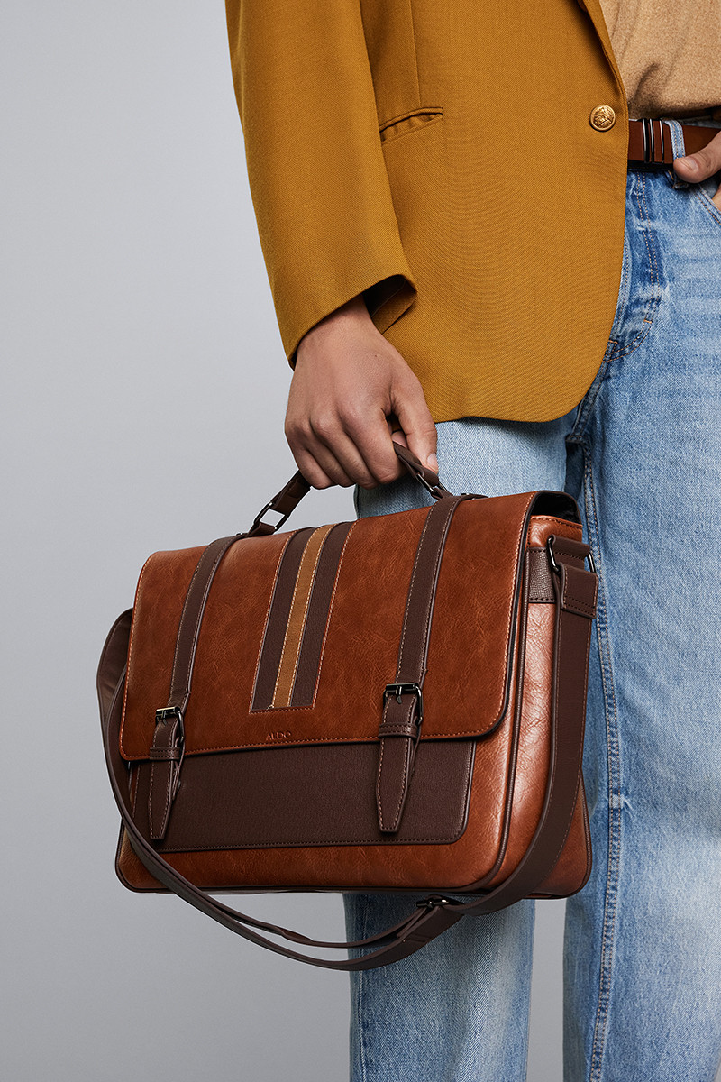 Man holding a stylish brown leather messenger bag, wearing a mustard blazer and blue jeans.