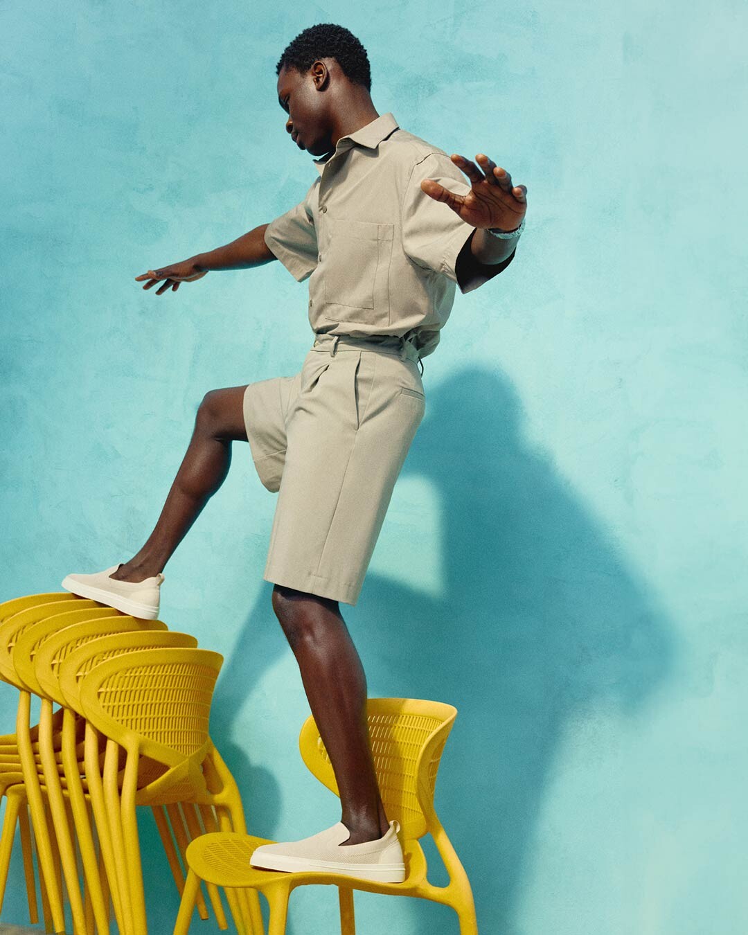 afro-descendant male model wearing beige espadrilles balancing himself on a pile of yellow chairs