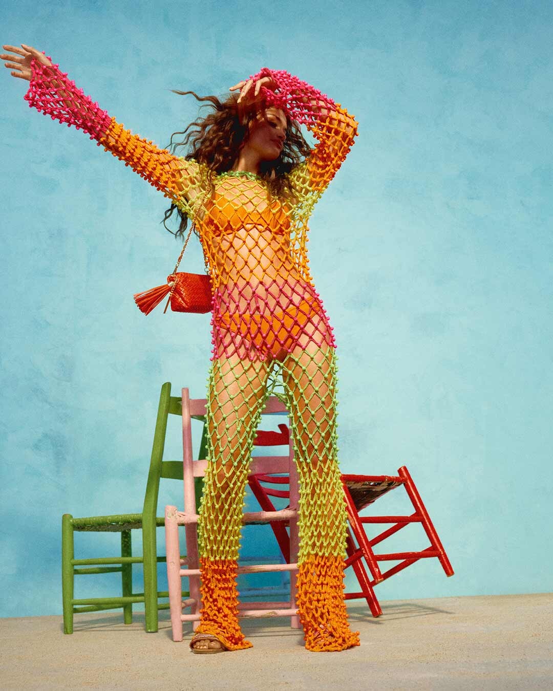 curly-haired girl dancing with her hands in the air wearing a colorful outfit and flat sandals next to 3 chairs and a blue wall