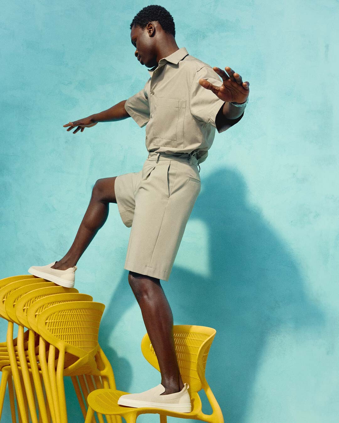 male model dressed in light colors and espadrilles balancing on yellow chairs