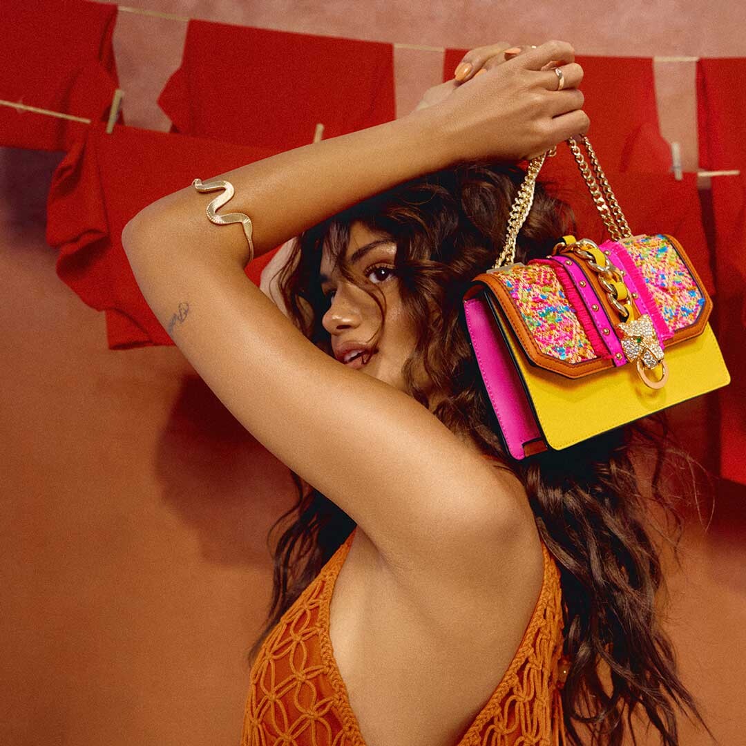 tanned woman in front of an orange wall with red clothes on a clothesline holding a colorful handbag to her head