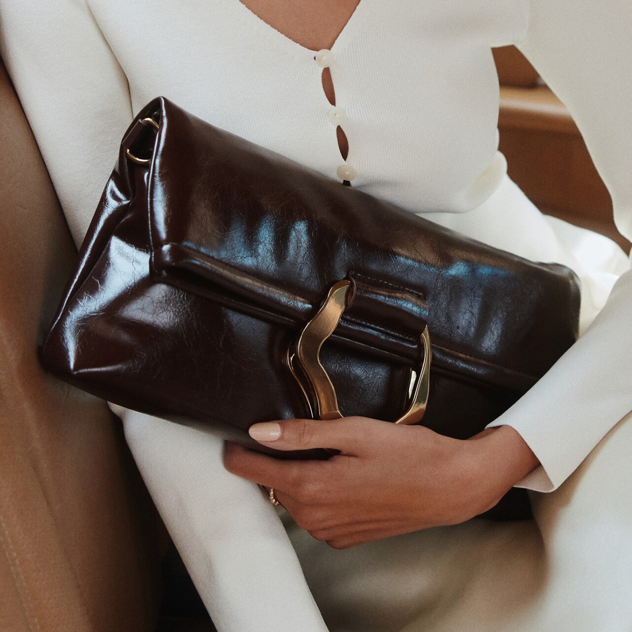 Mulher com roupa branca a segurar uma clutch castanha brilhante com fivela.