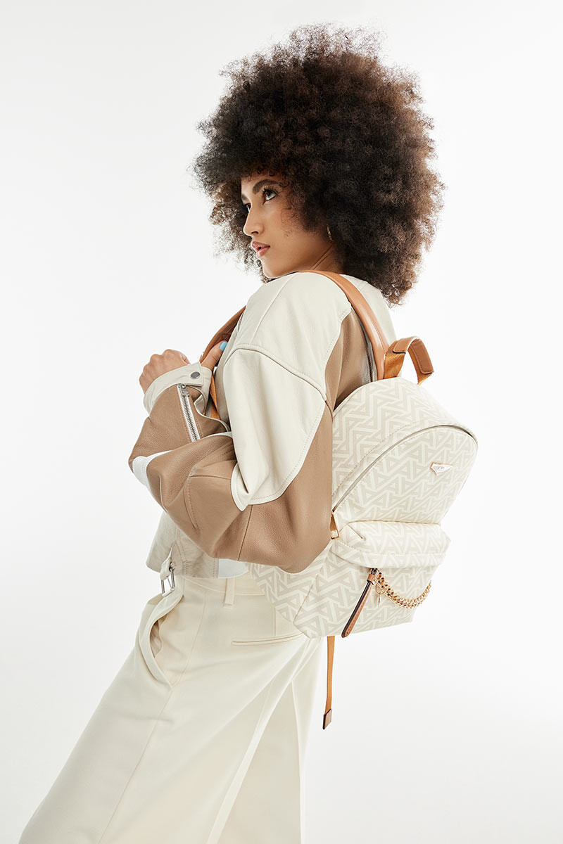 Mulher de cabelo afro de lado a usar roupas em cores bege a carregar uma mochila bege às costas com padrão ALDO e detalhes dourados