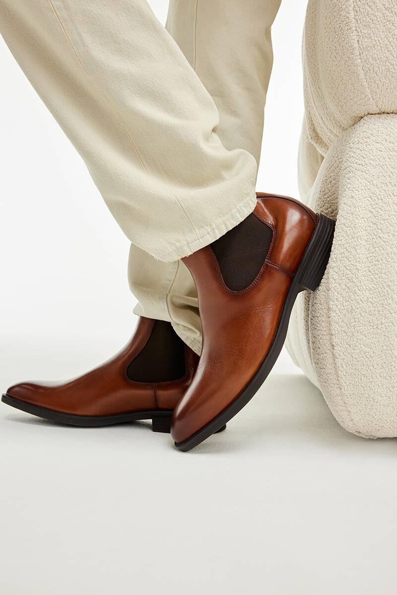 Pormenor da perna de homem junto a um sofá bege a usar calças de ganga bege e botins castanhos em pele com elástico