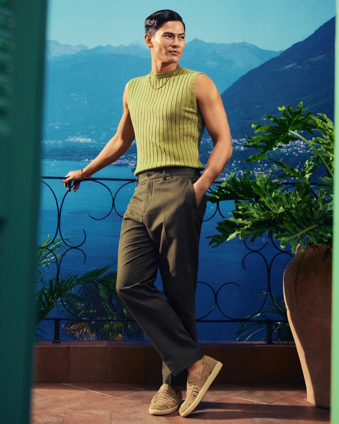 Un hombre de pie, vestido de verde con alpargatas ALDO, apoyado en un balcón con vistas al río.
