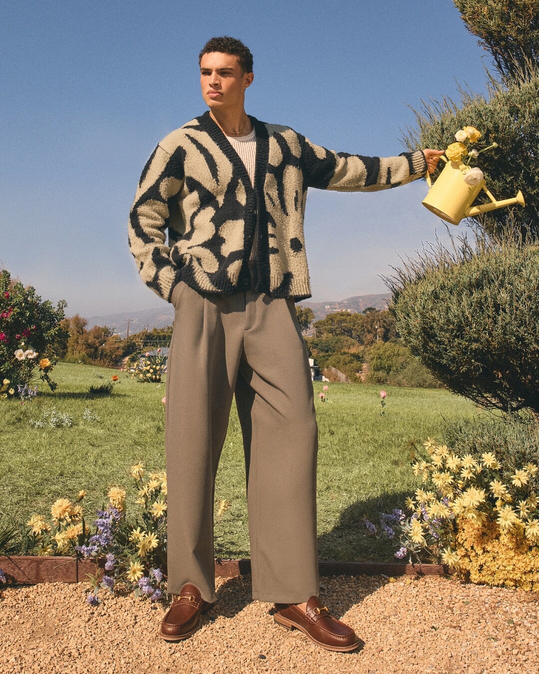 Homem num campo verde com flores, a segurar um regador amarelo e a usar uns mocassins castanhos.