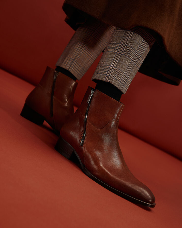 Pormenor de pés de homem num fundo vermelho a usar botins castanhos em pele e fecho lateral, meia preta e calça formal com padrão xadrez 