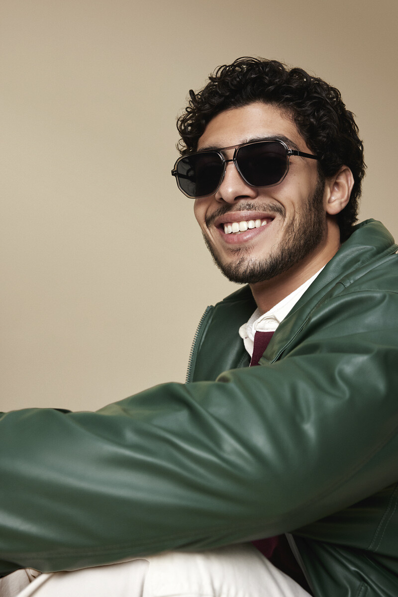 Homem a sorrir, sentado com uma casaco verde oversize e a usar óculos de sol escuros e com as lentes escuras também. 