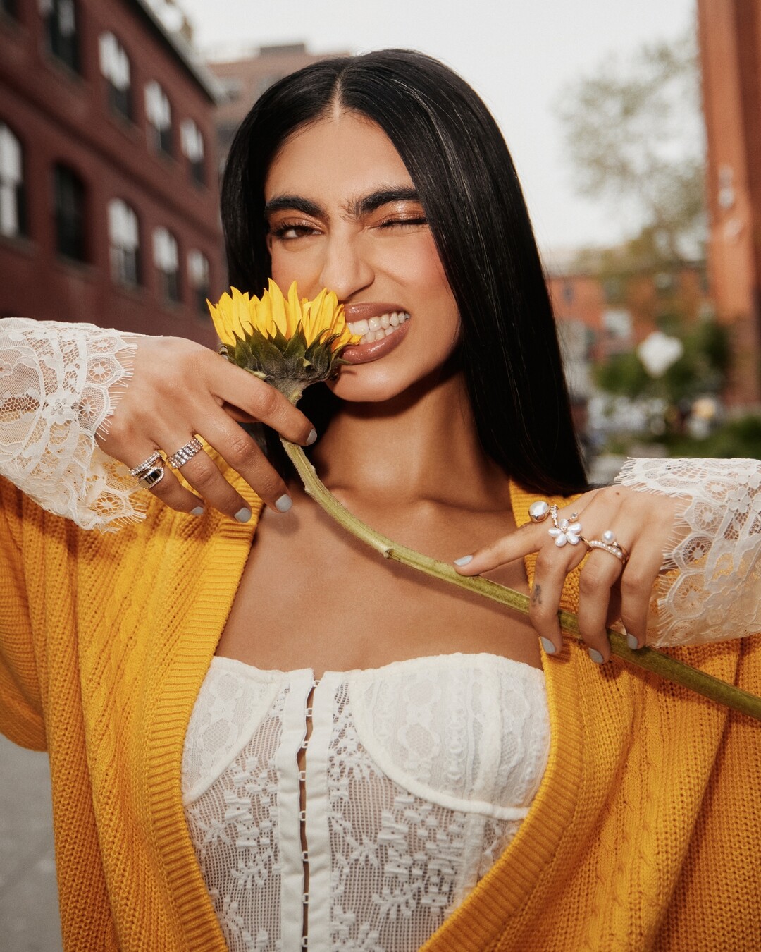 Mujer sonriendo alegremente mientras sostiene un girasol entre los dientes, vestida con una chaqueta amarilla y un top de encaje blanco, en un entorno urbano con edificios al fondo.