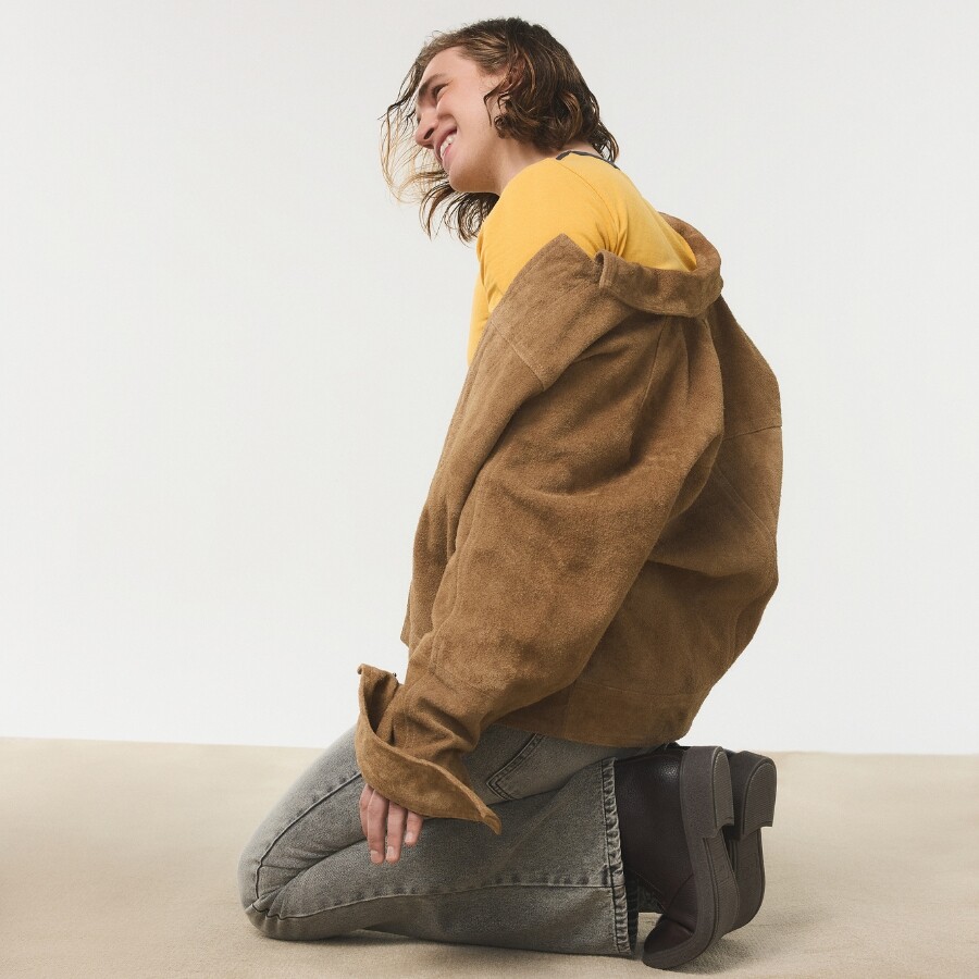 Joven arrodillado en el suelo, con chaqueta oversize camel y vaqueros grises, llevando zapatos de plataforma negros.