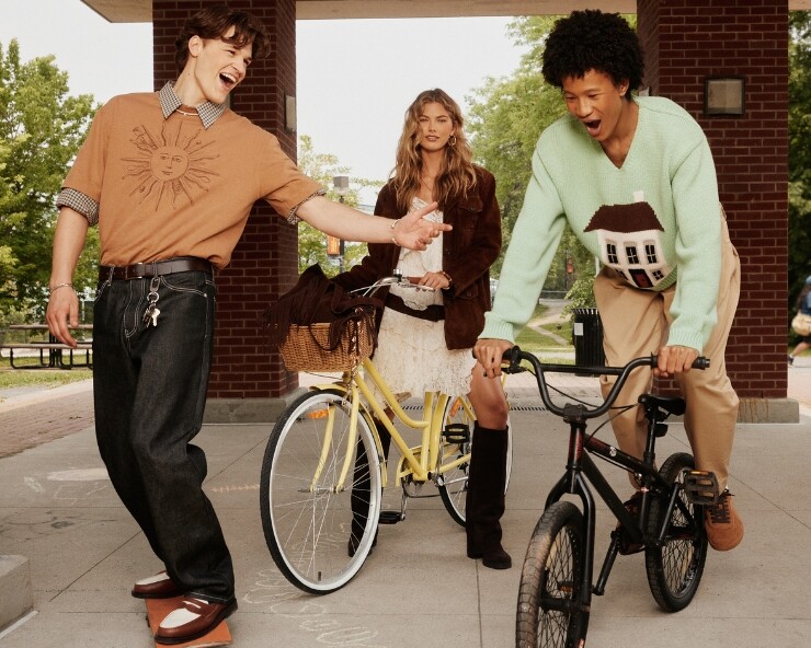 Tres jóvenes con looks modernos y desenfadados se divierten al aire libre: dos chicos con bicicletas y una chica en el centro con un vestido blanco y una bicicleta amarilla.