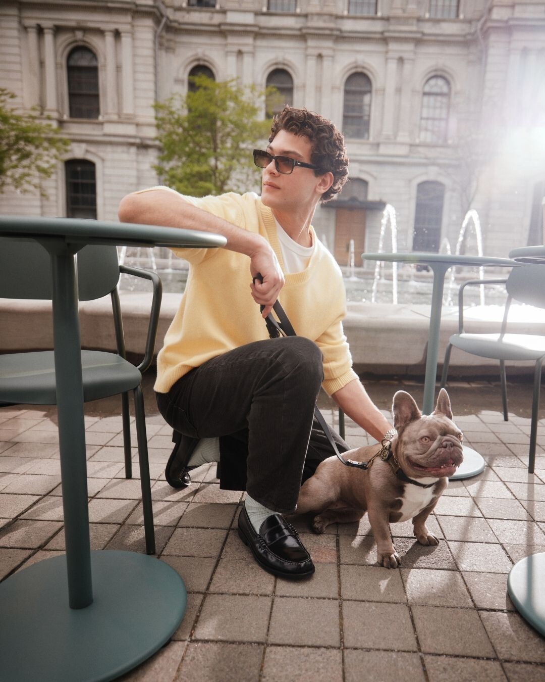 Hombre sentado en una terraza con bulldog francés, con jersey amarillo, pantalón de pana negro y mocasines de piel negra, colección FW26 ALDO.