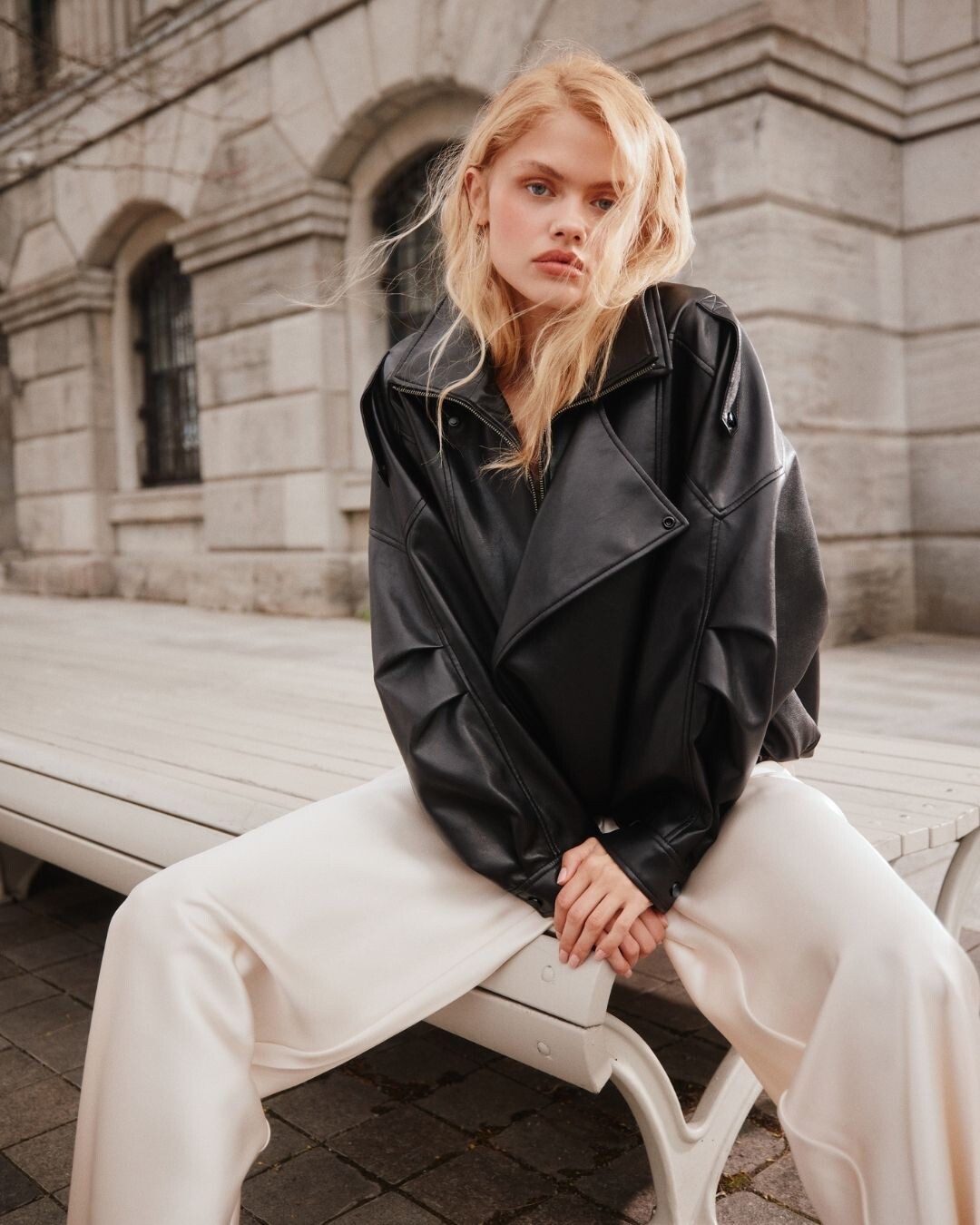 Mujer rubia sentada en un banco, con chaqueta de piel negra oversized y pantalón blanco de sastrería, colección FW26 ALDO.