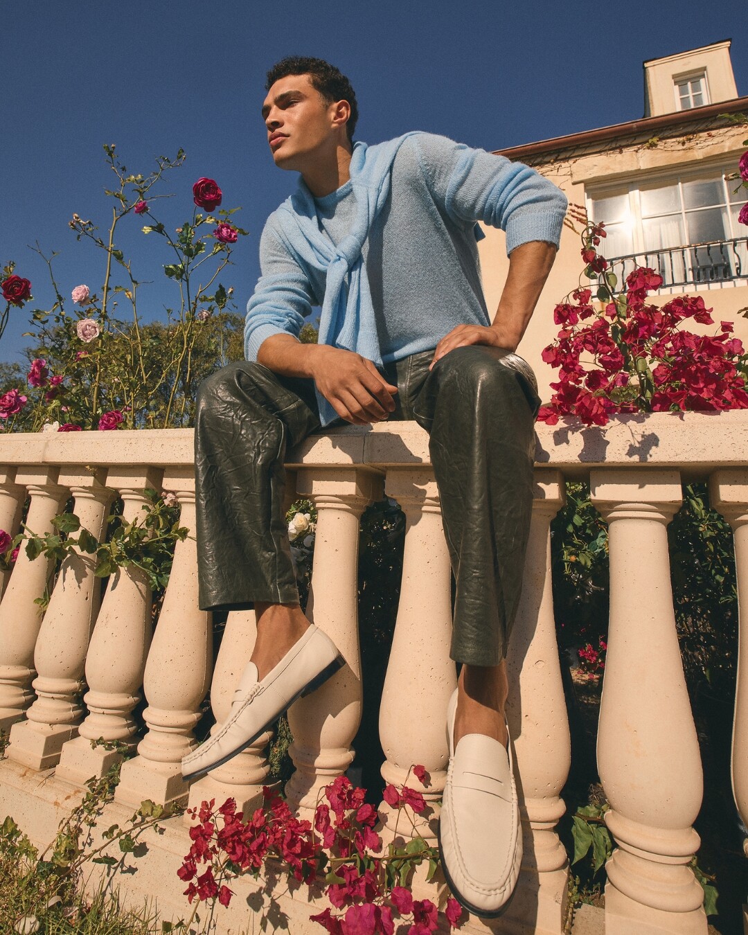 Homem com camisola azul, calças pretas e mocassins brancos está sentado num corrimão ao sol, rodeado de flores cor-de-rosa
