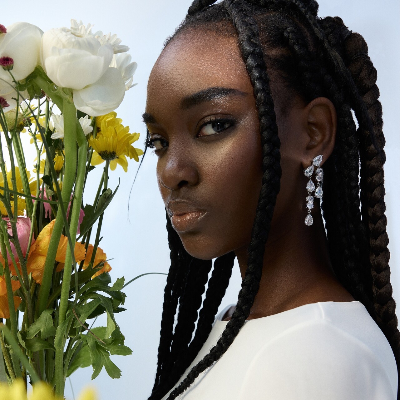 Mujer con trenzas y pendientes brillantes junto a flores.