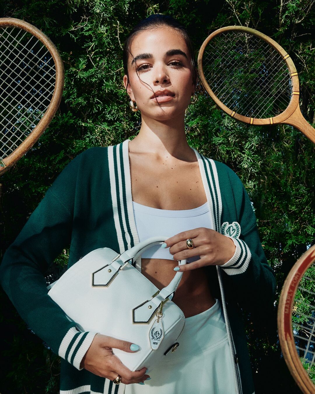 Uma mulher com um casaco desportivo verde apresenta-se confiante a segurar numa mala branca.