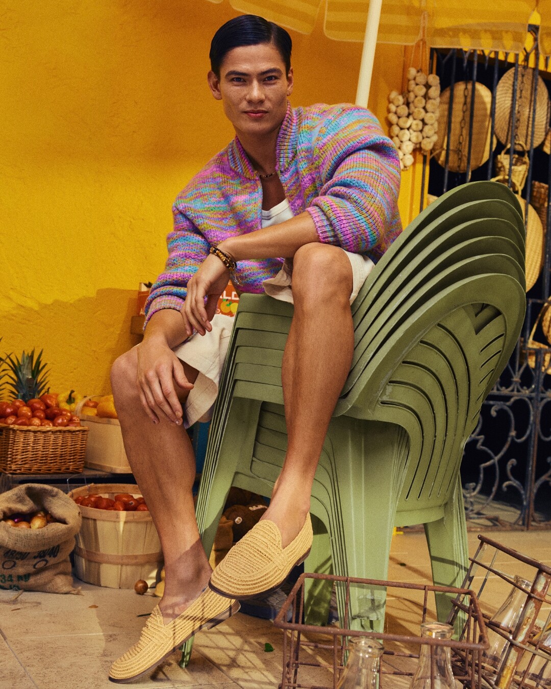 Homem vestido com casaco colorido, calções brancos e sapatos de ráfia da ALDO, sentado em cima de cadeiras verdes empilhadas. 