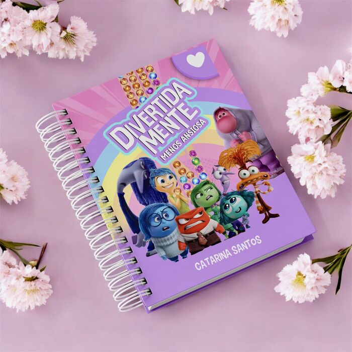Um caderno com espiral sobre um fundo rosa com flores brancas. A capa traz personagens de desenhos animados de DivertidaMente e o texto 