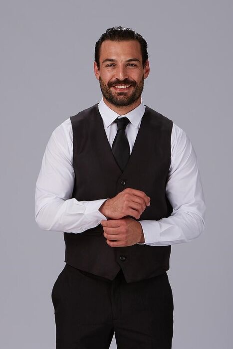 Homem com colete preto, camisa branca e gravata preta, sorrindo e a ajustar a manga.