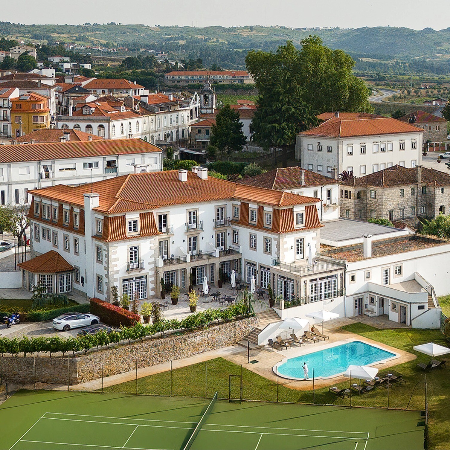 Vista aérea do Forrester Essence Douro Hotel em Alijó, destacando o palacete histórico com telhados vermelhos, piscina exterior, campos de ténis e a envolvente paisagem da vila e das colinas do Douro.