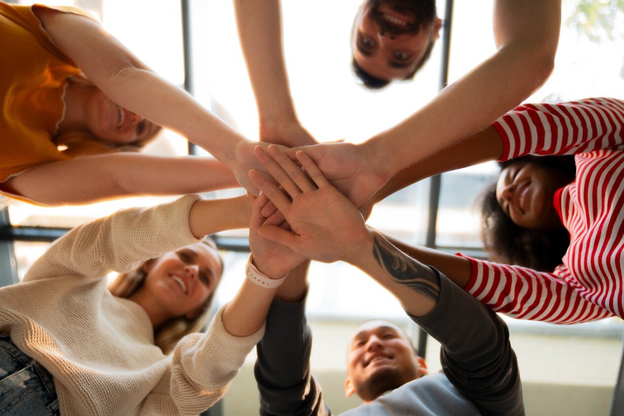 Grupo de pessoas a colocar as mãos juntas em sinal de união e trabalho em equipa, num ambiente alegre e descontraído.