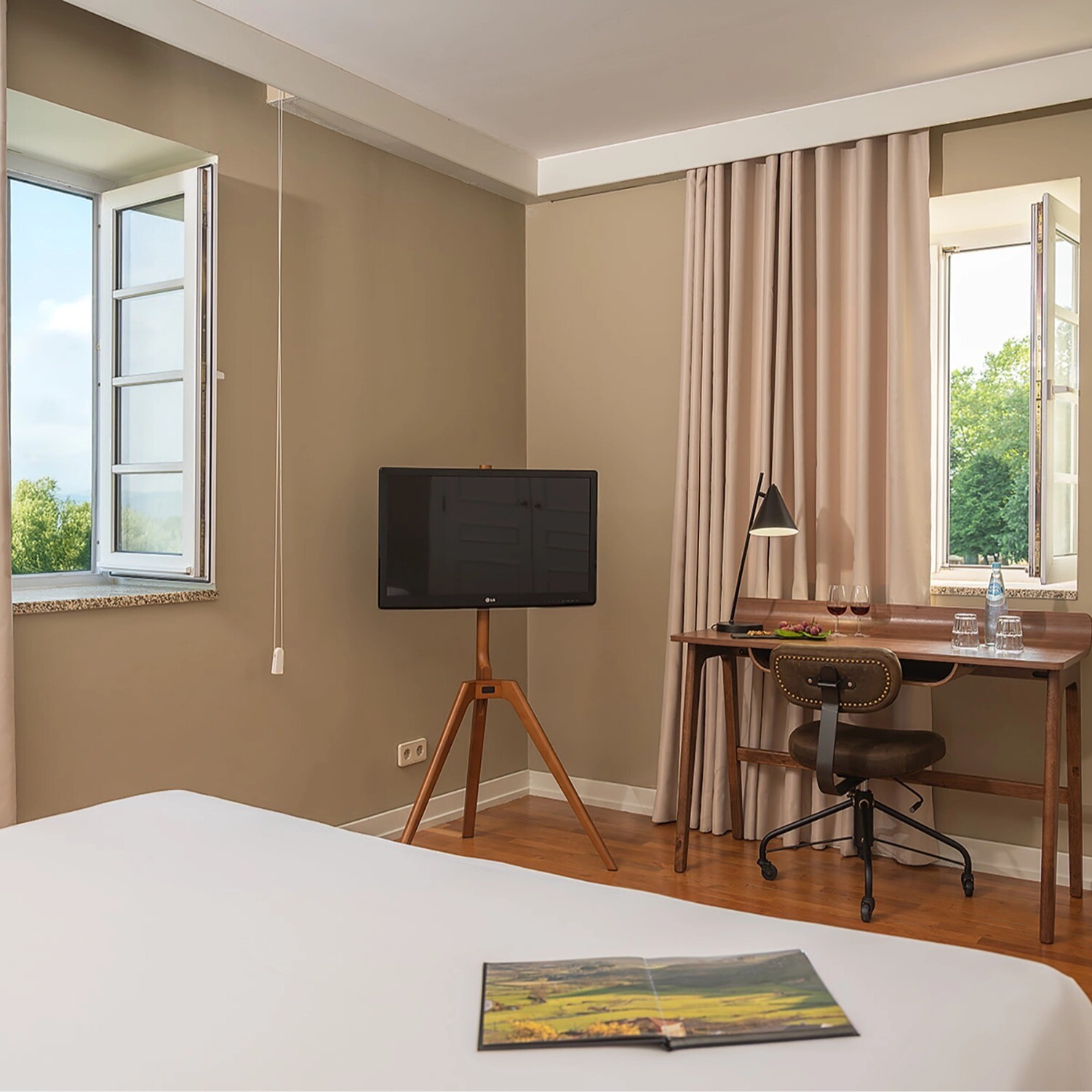 Quarto Prestige com janelas abertas, secretária, televisão e luz natural.
