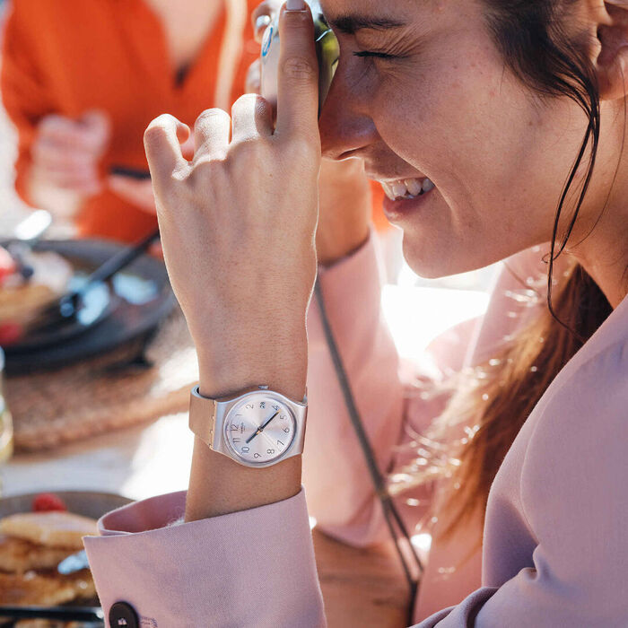 Modelo feminino com um relógio Swatch de bracelete rosa e um mostrador analógico com calendário.