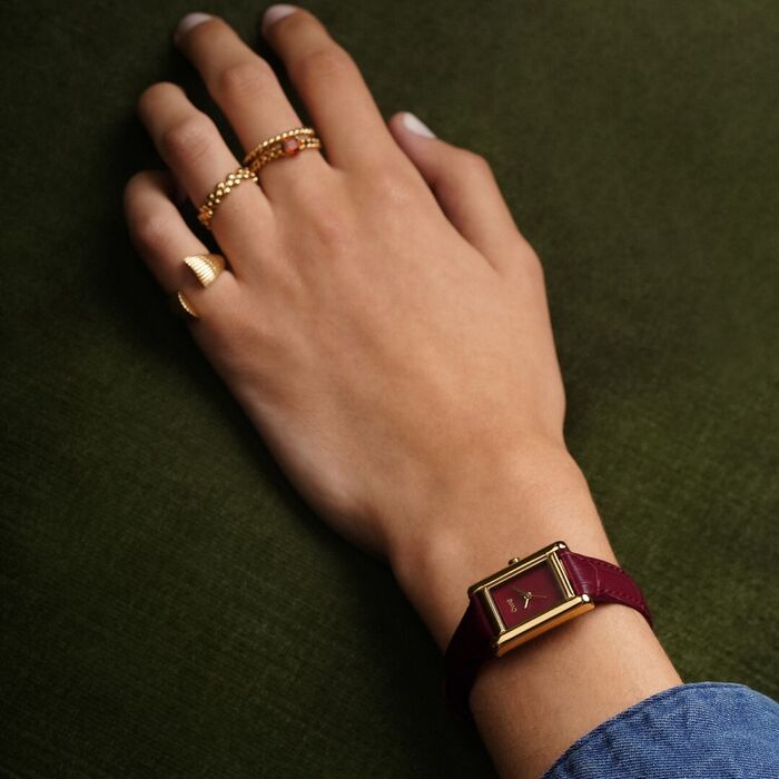 Mão de modelo com relógio One de bracelete em bordeaux e com caixa retangular dourada.