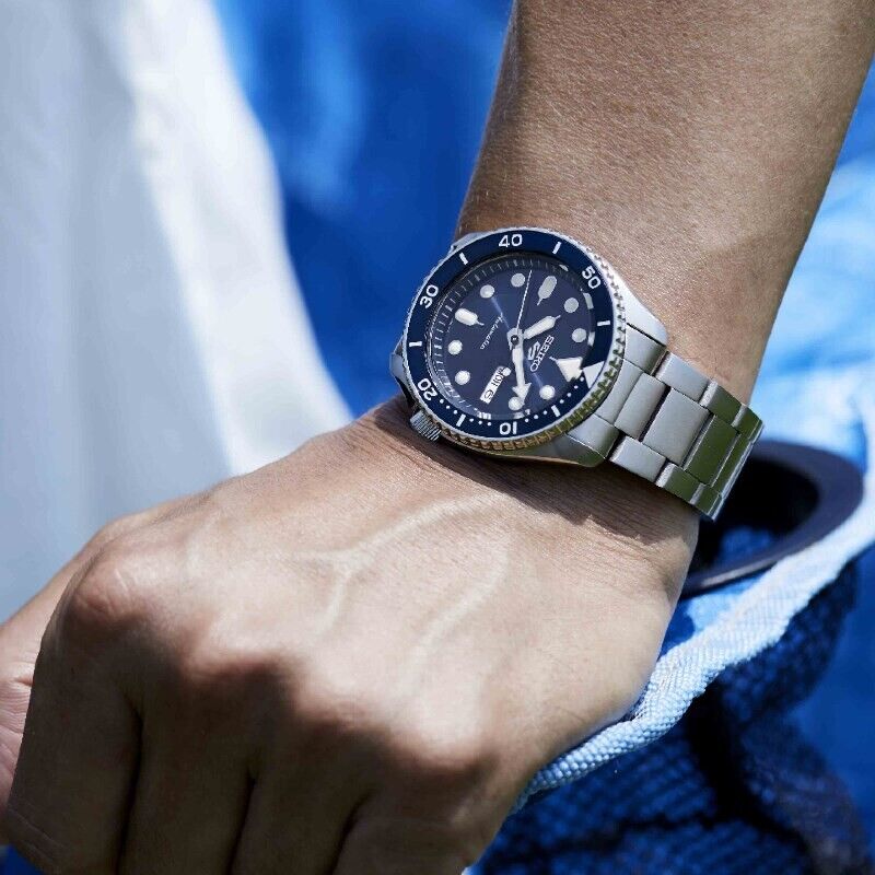 Pulso masculino com relógio Seiko 5 com bracelete e caixa em aço cor prateada e mostrador azul.