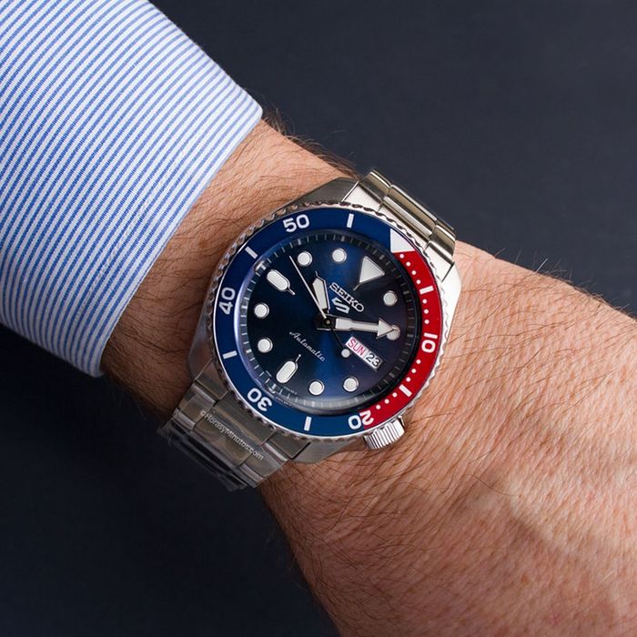 Pulso masculino com relógio Seiko 5 com bracelete e caixa em aço cor prateada e mostrador azul.