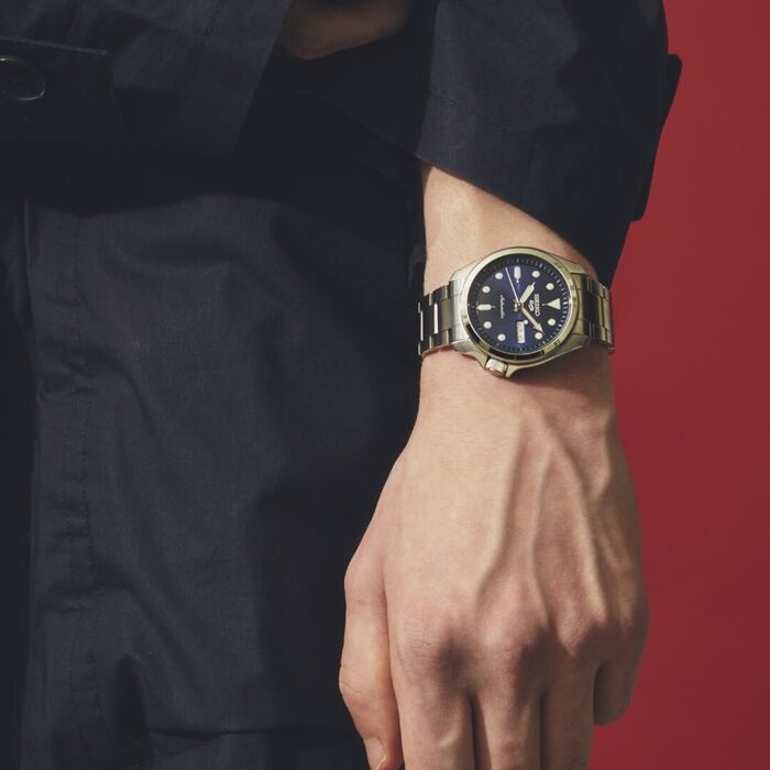 Pulso masculino com relógio Seiko 5, com bracelete e caixa em aço de cor prateada e mostrador azul.