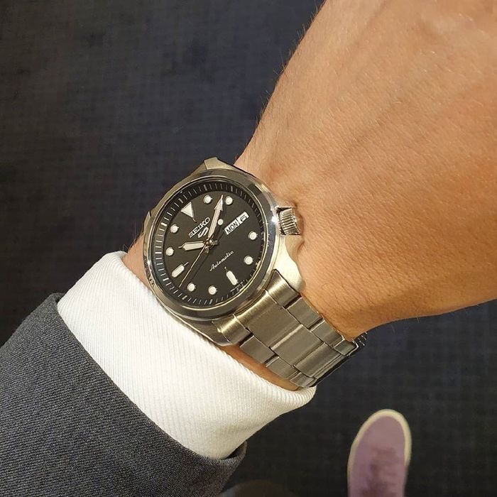 Pulso masculino com relógio Seiko 5, com bracelete e caixa em aço de cor prateada e mostrador preto.