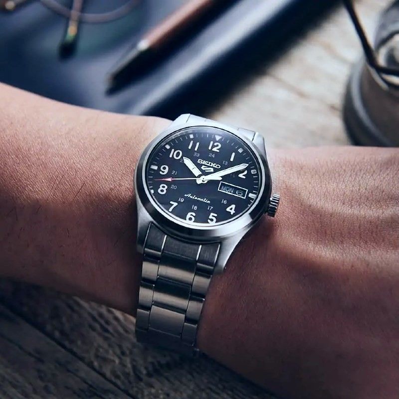 Pulso masculino com relógio Seiko 5, com bracelete e caixa em aço de cor prateada e mostrador azul.