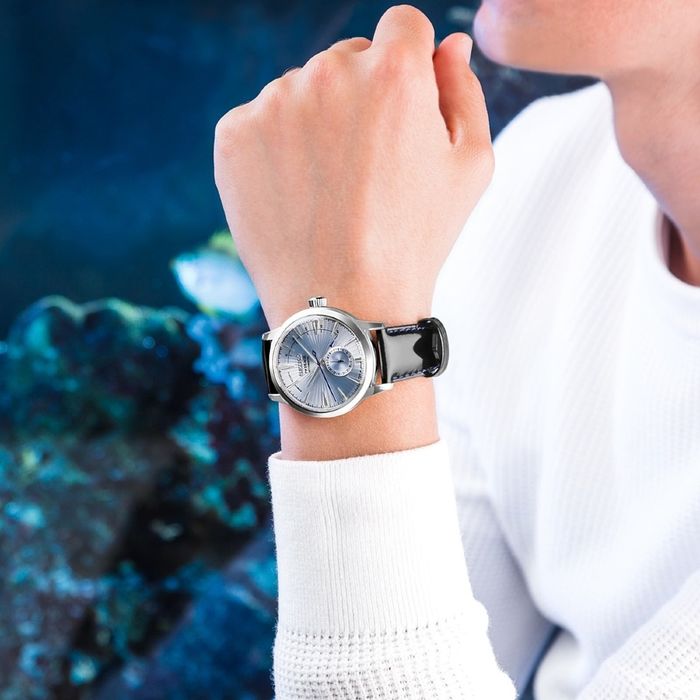 Pulso masculino com relógio Seiko, de bracelete em pele preta e caixa prateada com mostrador azul.