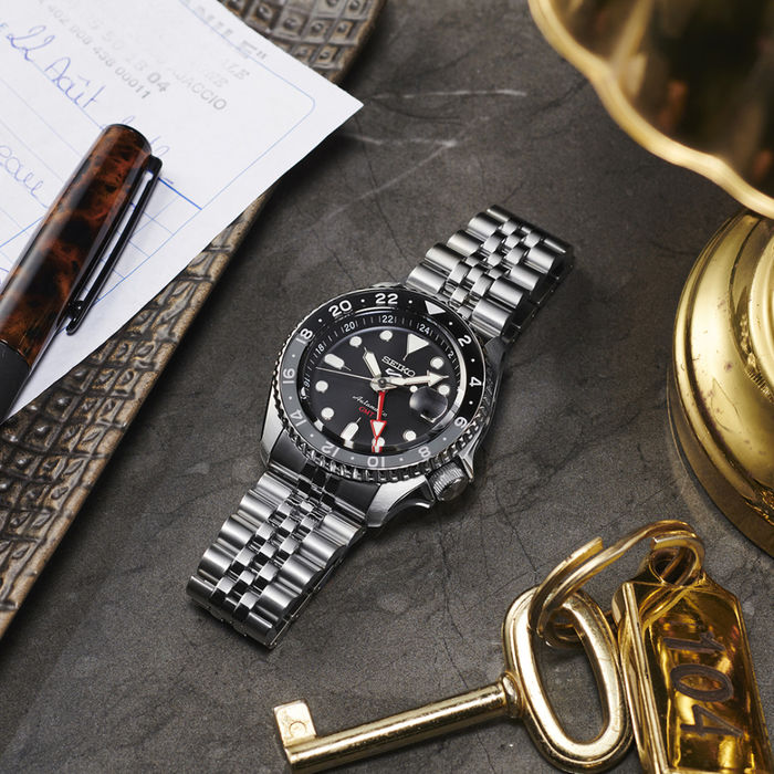 Modelo Seiko 5 de bracelete e caixa em aço prateado e mostrador preto, pousado em uma mesa preta.