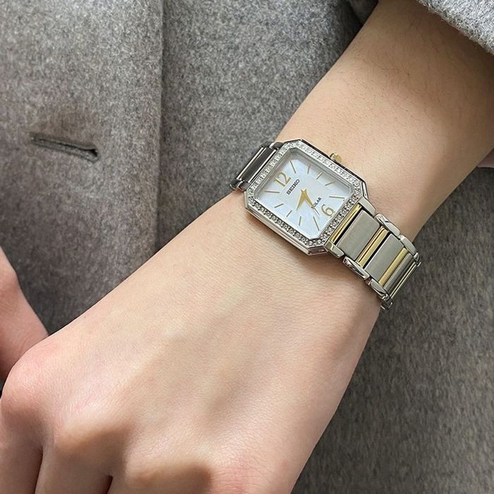 Pulso feminino com Seiko de bracelete e caixa prateada, mostrador retangular madrepérola e pedras.