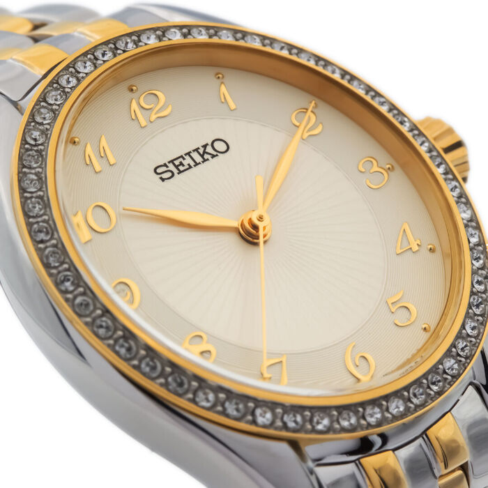Vista aproximada de mostrador Seiko feminino com pedras à volta do mostrador e cor champanhe.