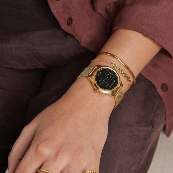 Pulso de senhora a usar smartwatch One, anéis e pulseiras da marca, de cor dourada