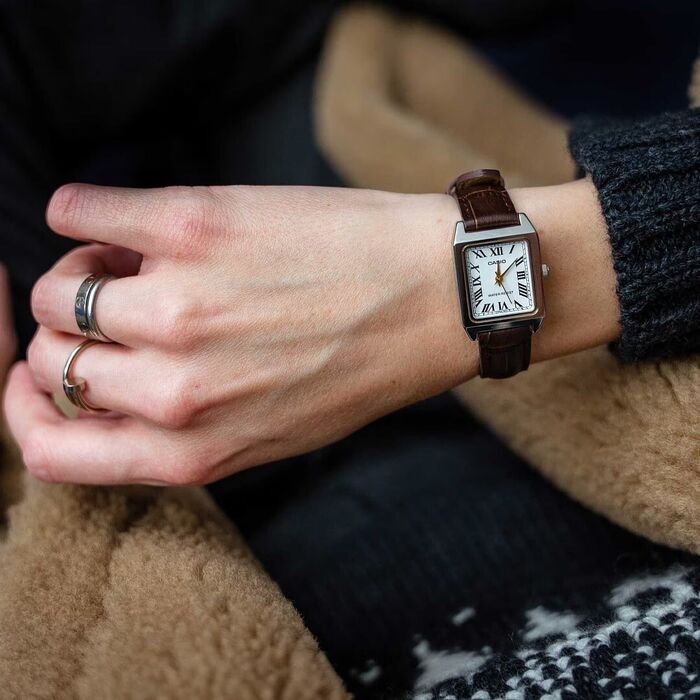 Pulso com relógio rectangular de mostrador branco com numerais romanos e bracelete castanha.