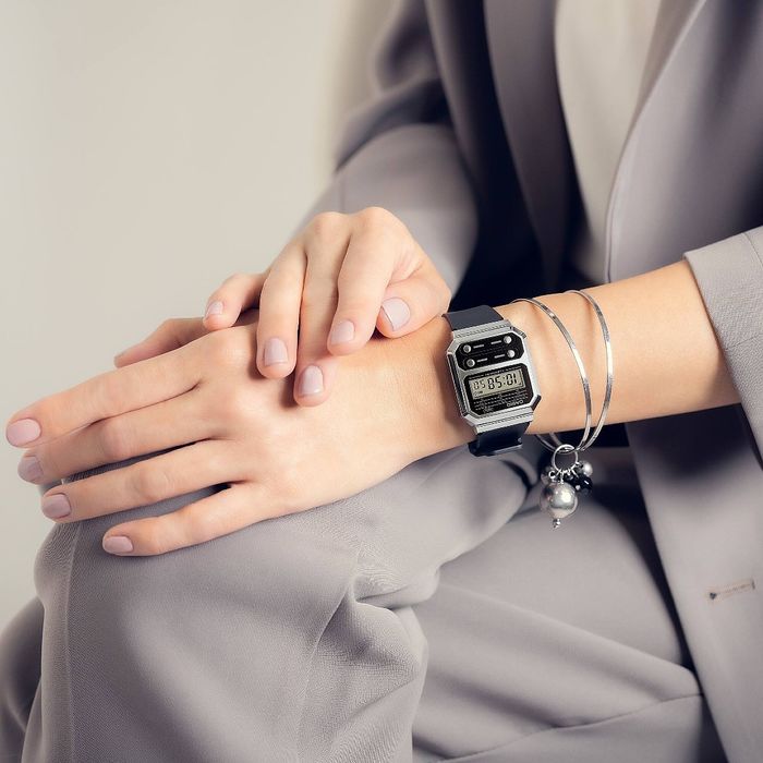 Mão de pessoa com fato cinzento a usar relógio digital preto com caixa prateada e pulseiras.