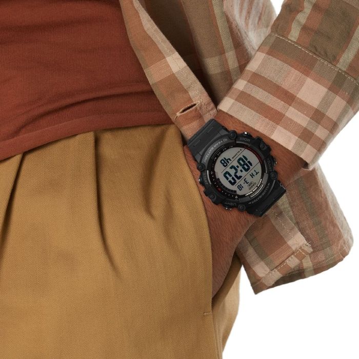 Pulso com relógio digital preto CASIO AE-1500WH e pessoa com camisa aos quadrados e calças castanhas.