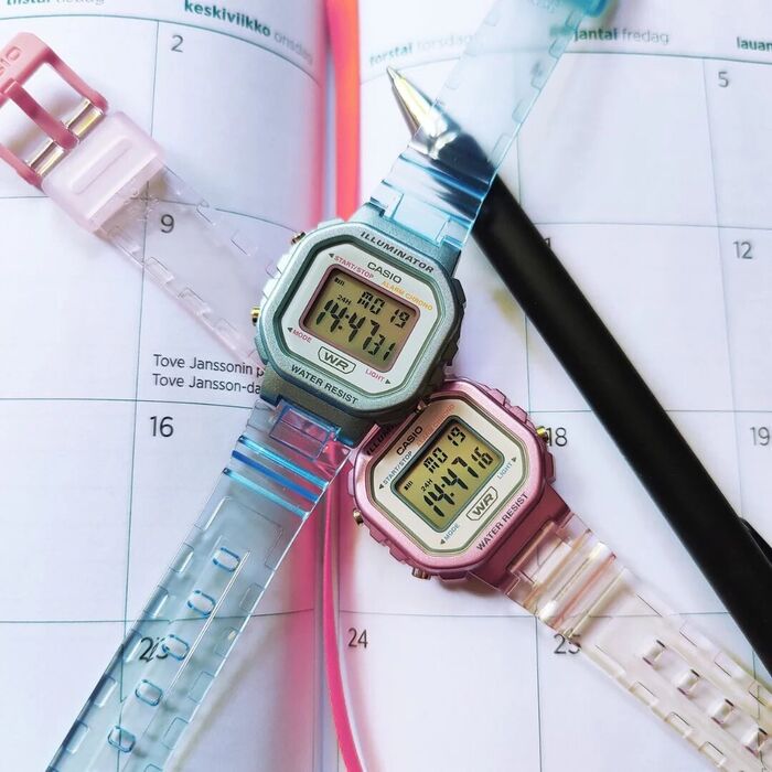 Dois relógios digitais com braceletes translúcidas azul e rosa sobre agenda com caneta.